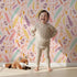 Lovely child standing in front of Whimsy Tots Feather Arrow Confetti Wallpaper in Petal Pink with arrow, feather, triangle, dot in design.