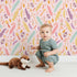 Lovely child standing in front of Whimsy Tots Feather Arrow Confetti Wallpaper in Blush Cream with arrow, feather, triangle, dot in design.