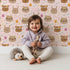 Lovely child standing in front of Whimsy Tots Feather Crown Critters Wallpaper in Petal Pink with bear, rabbit, crown, feather, arrow, dot in design.