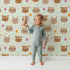Lovely child standing in front of Whimsy Tots Feather Crown Critters Wallpaper in Meadow Green with bear, rabbit, crown, feather, arrow, dot in design.