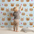 Lovely child standing in front of Whimsy Tots Feather Crown Critters Wallpaper in Soft Sky Blue with bear, rabbit, crown, feather, arrow, dot in design.