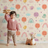 Lovely child standing in front of Whimsy Tots Birdie Rainbow Float Parade Wallpaper in Vanilla Glow with bird, balloon, rainbow, heart, cloud in design.