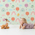 Lovely child standing in front of Whimsy Tots Birdie Rainbow Float Parade Wallpaper in Mint Daydream Green with bird, balloon, rainbow, heart, cloud in design.