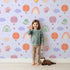 Lovely child standing in front of Whimsy Tots Birdie Rainbow Float Parade Wallpaper in Lavender Daydream with bird, balloon, rainbow, heart, cloud in design.