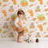 Lovely child standing in front of Whimsy Tots Catnap and Chirp Parade Wallpaper in Cream with cat, bird, box, heart, confetti in design.