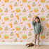 Lovely child standing in front of Whimsy Tots Catnap and Chirp Parade Wallpaper in Blush Pink with cat, bird, box, heart, confetti in design.