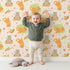 Lovely child standing in front of Whimsy Tots Catnap and Chirp Parade Wallpaper in Beige with cat, bird, box, heart, confetti in design.