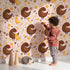 Lovely child standing in front of Whimsy Tots Hen and Chick Meadow Wallpaper in Blush Nest Pink with chicken, chick, flower, leaf in design.