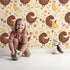Lovely child standing in front of Whimsy Tots Hen and Chick Meadow Wallpaper in Honey Wheat Yellow with chicken, chick, flower, leaf in design.