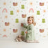 Lovely child standing in front of Whimsy Tots Garden Hop and Snail Trail Wallpaper in White with frog, snail, dragonfly, rainbow, sun, flower in design.
