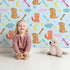 Lovely child standing in front of Whimsy Tots Arrow Pup and Foxy Night Parade Wallpaper in Sky Blue with dog, fox, moon, arrow, star, cloud in design.