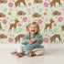 Lovely child standing in front of Whimsy Tots Blossom Meadow Deer Wallpaper in Mint Green with deer, flower, butterfly, heart in design.