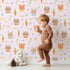Lovely child standing in front of Whimsy Tots Meadow Crown Friends Wallpaper in Blush Pink with fox, rabbit, bear, flower, carrot, crown, bow in design.