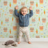 Lovely child standing in front of Whimsy Tots Meadow Crown Friends Wallpaper in Mint Green with fox, rabbit, bear, flower, carrot, crown, bow in design.