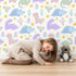 Lovely child standing in front of Whimsy Tots Cactus Dino Parade Wallpaper in White with dinosaur, cactus, flower in design.