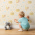 Lovely child standing in front of Whimsy Tots Snail Meadow Cuddle Parade Wallpaper in Cream with snail, flower, leaf in design.