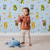 Lovely child standing in front of Whimsy Tots Wild Heart Animal Parade Wallpaper in Sky Blue with giraffe, koala, leopard, monkey, tiger, crocodile, heart, arrow, star in design.
