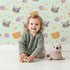 Lovely child standing in front of Whimsy Tots Meadow Snuggle Faces Wallpaper in Mint Green with cat, raccoon, flower, heart in design.