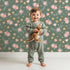 Lovely child standing in front of Whimsy Tots Twilight Wildflower Waltz Wallpaper in Forest Green with flower, star, leaf in design.