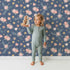 Lovely child standing in front of Whimsy Tots Twilight Wildflower Waltz Wallpaper in Blue with flower, star, leaf in design.
