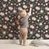 Lovely child standing in front of Whimsy Tots Twilight Wildflower Waltz Wallpaper in Charcoal Gray with flower, star, leaf in design.