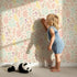 Lovely child standing in front of Whimsy Tots Whimsy Meadow Sketch Wallpaper in Cream with leaf, flower, branch in design.