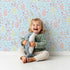Lovely child standing in front of Whimsy Tots Whimsy Meadow Sketch Wallpaper in Sky with leaf, flower, branch in design.