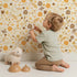 Lovely child standing in front of Whimsy Tots Golden Woodland Whimsy Wallpaper in Cream with leaf, flower, berry, crown in design.