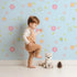 Lovely child standing in front of Whimsy Tots Breezy Blossom Meadow Wallpaper in Sky with flower, petal, leaf in design.