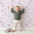 Lovely child standing in front of Whimsy Tots Fairy Petal Garden Wallpaper in Pink with fairy, flower, leaf, berry in design.