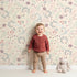 Lovely child standing in front of Whimsy Tots Fairy Petal Garden Wallpaper in Cream with fairy, flower, leaf, berry in design.