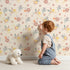 Lovely child standing in front of Whimsy Tots Sunbeam Wildflower Parade Wallpaper in Beige with flower, wildflower, leaf, bouquet in design.