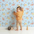 Lovely child standing in front of Whimsy Tots Sunbeam Wildflower Parade Wallpaper in Blue with flower, wildflower, leaf, bouquet in design.