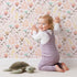 Lovely child standing in front of Whimsy Tots Blooming Meadow Stems Wallpaper in Blush Pink with flower, leaf, wildflower, stem in design.