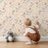 Lovely child standing in front of Whimsy Tots Blooming Meadow Stems Wallpaper in Beige with flower, leaf, wildflower, stem in design.