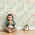 Lovely child standing in front of Whimsy Tots Blooming Meadow Stems Wallpaper in Mint Green with flower, leaf, wildflower, stem in design.