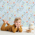Lovely child standing in front of Whimsy Tots Blooming Meadow Stems Wallpaper in Blue with flower, leaf, wildflower, stem in design.