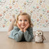 Lovely child standing in front of Whimsy Tots Blooming Meadow Stems Wallpaper in White with flower, leaf, wildflower, stem in design.