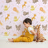 Lovely child standing in front of Whimsy Tots Safari Buddies Parade Wallpaper in Pink with lion, giraffe, elephant, hippo, tiger, arrow in design.
