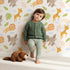 Lovely child standing in front of Whimsy Tots Jungle Leaf Pals Wallpaper in Cream with lion, giraffe, elephant, leaf, cloud in design.