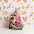 Lovely child standing in front of Whimsy Tots Jungle Leaf Pals Wallpaper in Blush Pink with lion, giraffe, elephant, leaf, cloud in design.