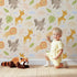 Lovely child standing in front of Whimsy Tots Jungle Leaf Pals Wallpaper in Sand with lion, giraffe, elephant, leaf, cloud in design.