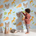 Lovely child standing in front of Whimsy Tots Jungle Leaf Pals Wallpaper in Blue with lion, giraffe, elephant, leaf, cloud in design.