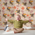 Lovely child standing in front of Whimsy Tots Tropical Toucan Friends Wallpaper in Cream with toucan, flower, leaf, hibiscus in design.
