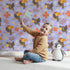 Lovely child standing in front of Whimsy Tots Tropical Toucan Friends Wallpaper in Lavender with toucan, flower, leaf, hibiscus in design.