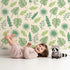 Lovely child standing in front of Whimsy Tots Tropical Leaf Mix Wallpaper in Cream with leaf, monstera, palm, jungle in design.