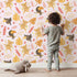 Lovely child standing in front of Whimsy Tots Monkey Banana Jungle Wallpaper in Pink with monkey, toucan, banana, leaf, flower in design.