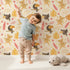 Lovely child standing in front of Whimsy Tots Monkey Banana Jungle Wallpaper in Cream with monkey, toucan, banana, leaf, flower in design.