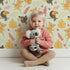 Lovely child standing in front of Whimsy Tots Monkey Banana Jungle Wallpaper in Mint Green with monkey, toucan, banana, leaf, flower in design.