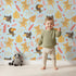 Lovely child standing in front of Whimsy Tots Monkey Banana Jungle Wallpaper in Sky Blue with monkey, toucan, banana, leaf, flower in design.